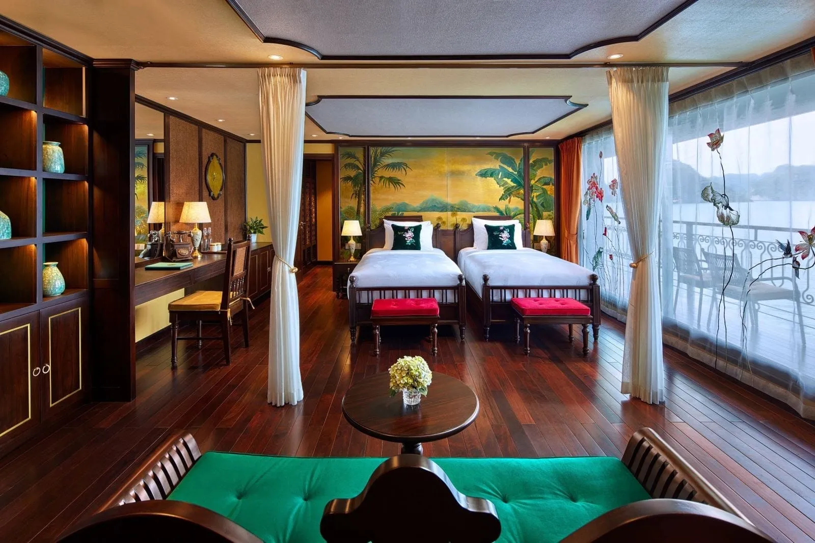 A wood-floored cabin with two twin beds with red benches, a desk, shelves with teal vases, a green daybed, and floor-to-ceiling sheer curtains opening onto a balcony with a view of a misty bay and karst hills.