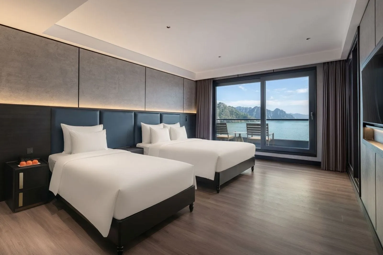 A modern cabin with two twin beds with white linens, dark headboards, wood floor, and a large window opening to a balcony with chairs and a view of the bay with limestone karst hills.