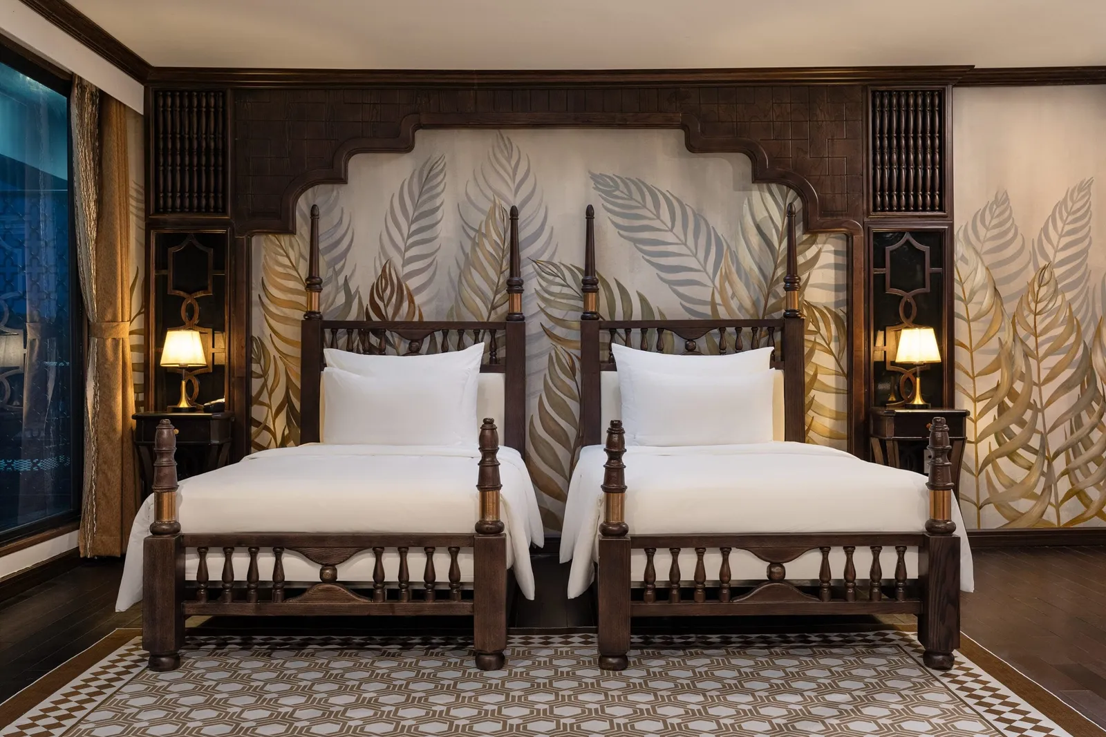 A cabin with two dark wood four-post twin beds with white linens, a leaf-patterned mural headboard wall, bedside lamps, a geometric rug and a window with gold curtains on the left.