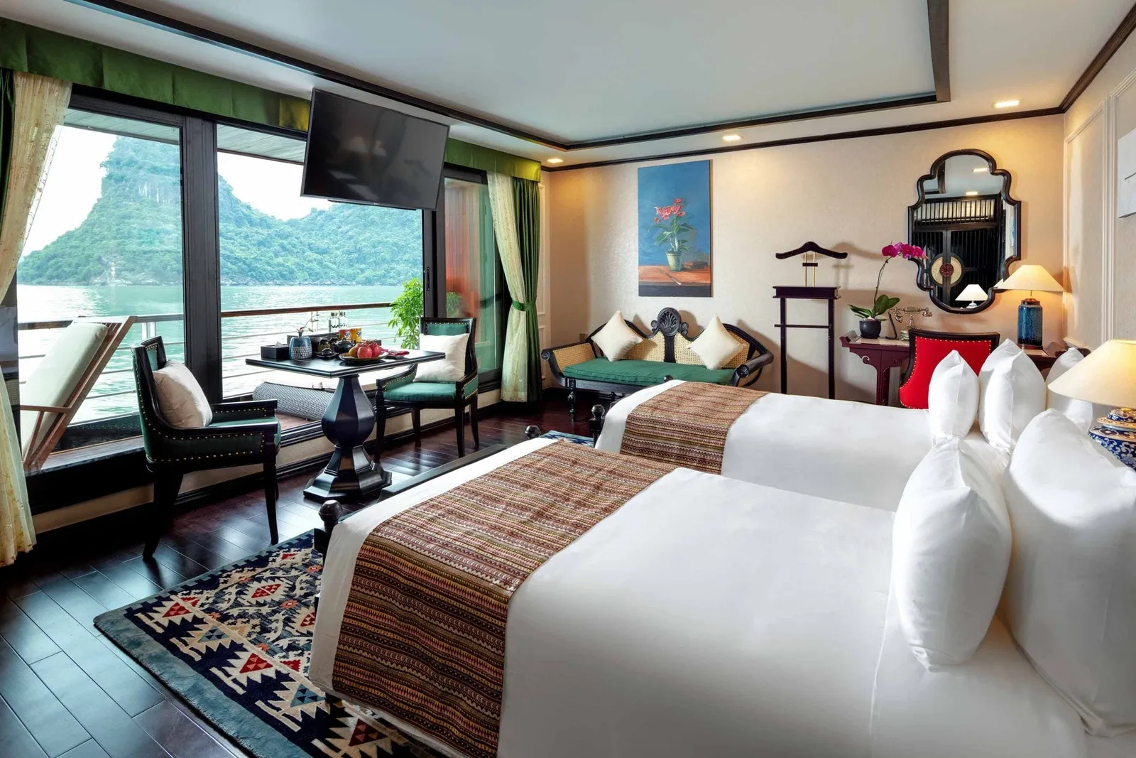A cabin with two twin beds, dark wood floor, green curtains and a table, with floor-to-ceiling windows and a balcony overlooking a bay with green karst hills.