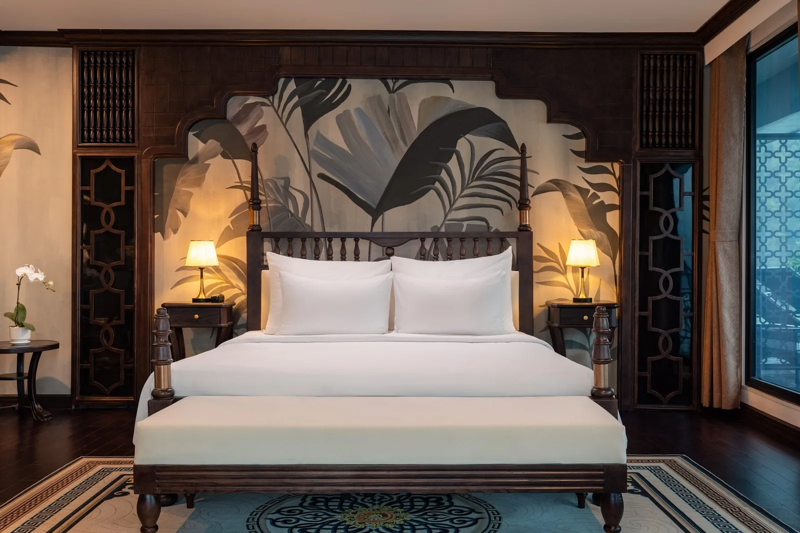 A luxury cabin interior with a dark wood four-poster bed, white bedding, an upholstered bench, a tropical leaf mural, two lit bedside lamps, an ornate patterned rug and a window on the right with a balcony.