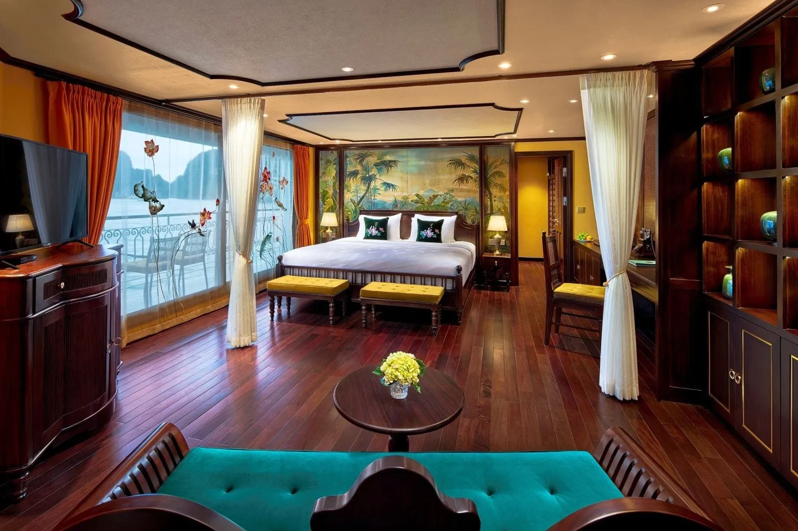 A spacious suite with dark hardwood floors, a king bed, a mural of palm trees, a teal sofa, and floor-to-ceiling windows showing a balcony and limestone karst silhouettes over water.