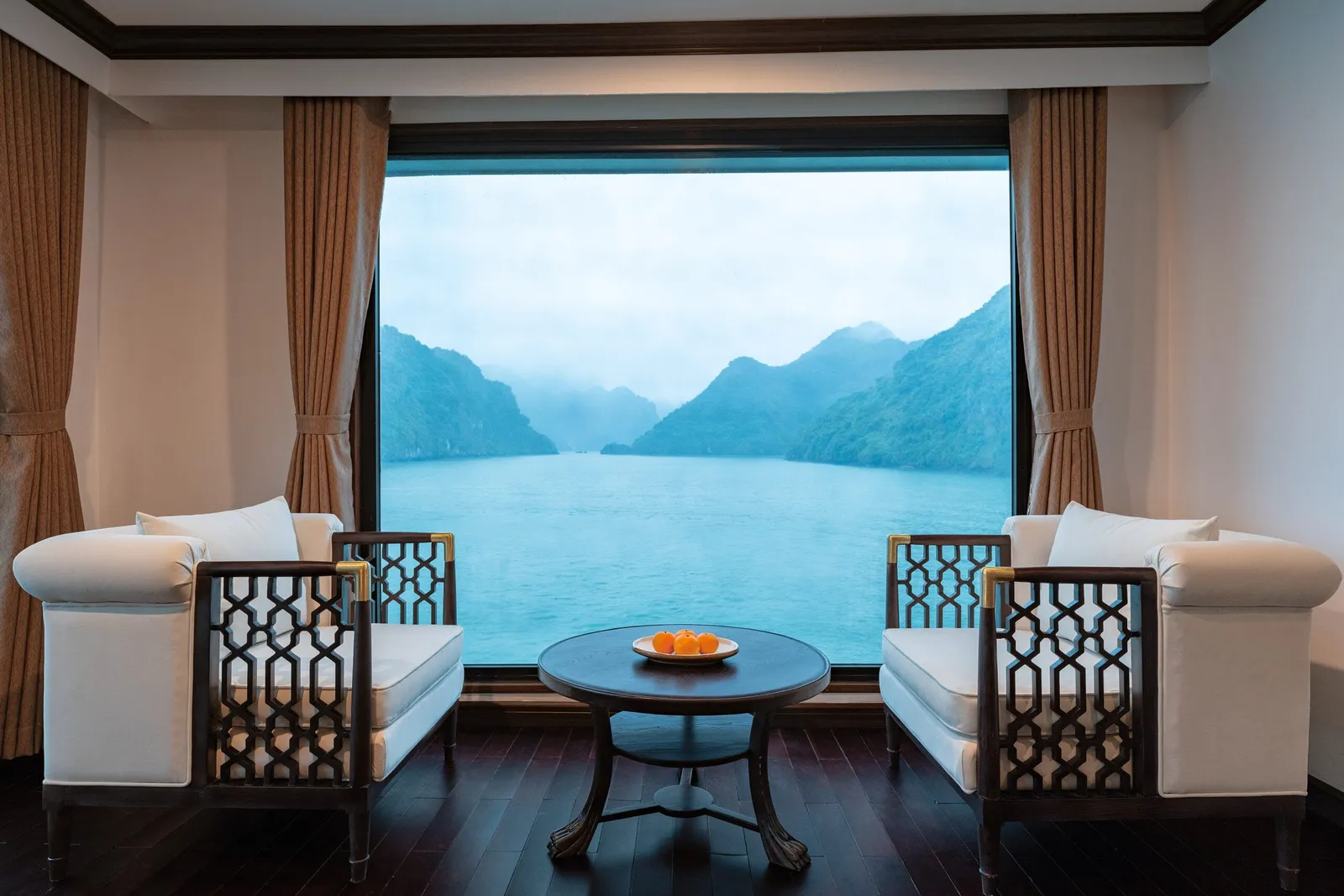 Interior lounge area with two white armchairs with dark lattice wood frames and a round table with oranges, facing a large window showing misty green karst islands over turquoise water.