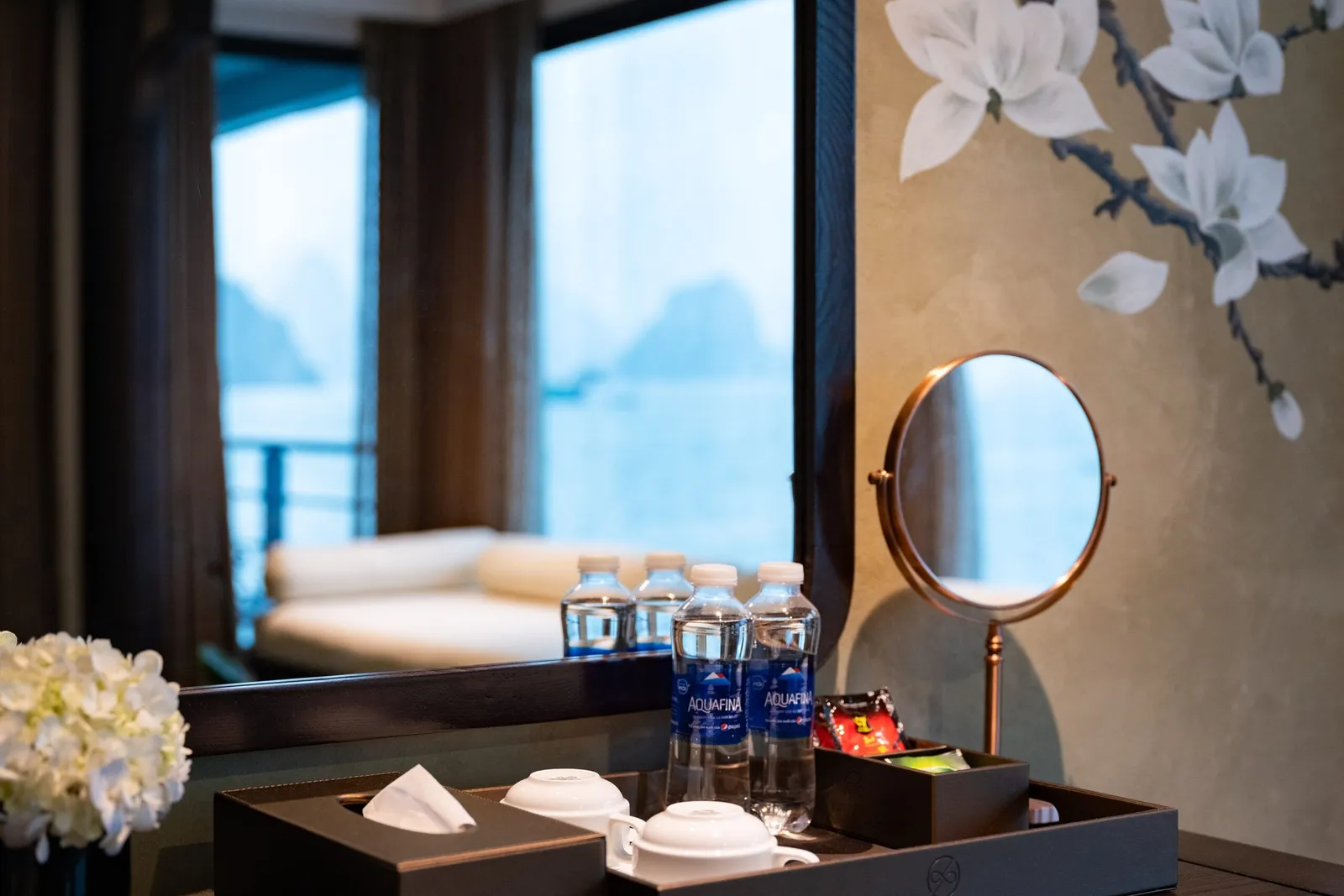 A cabin vanity with Aquafina water bottles, cups and a tissue box on a tray marked VENUS, a round mirror, white flowers, and a mirror reflecting a daybed and a window view of misty karst islands.