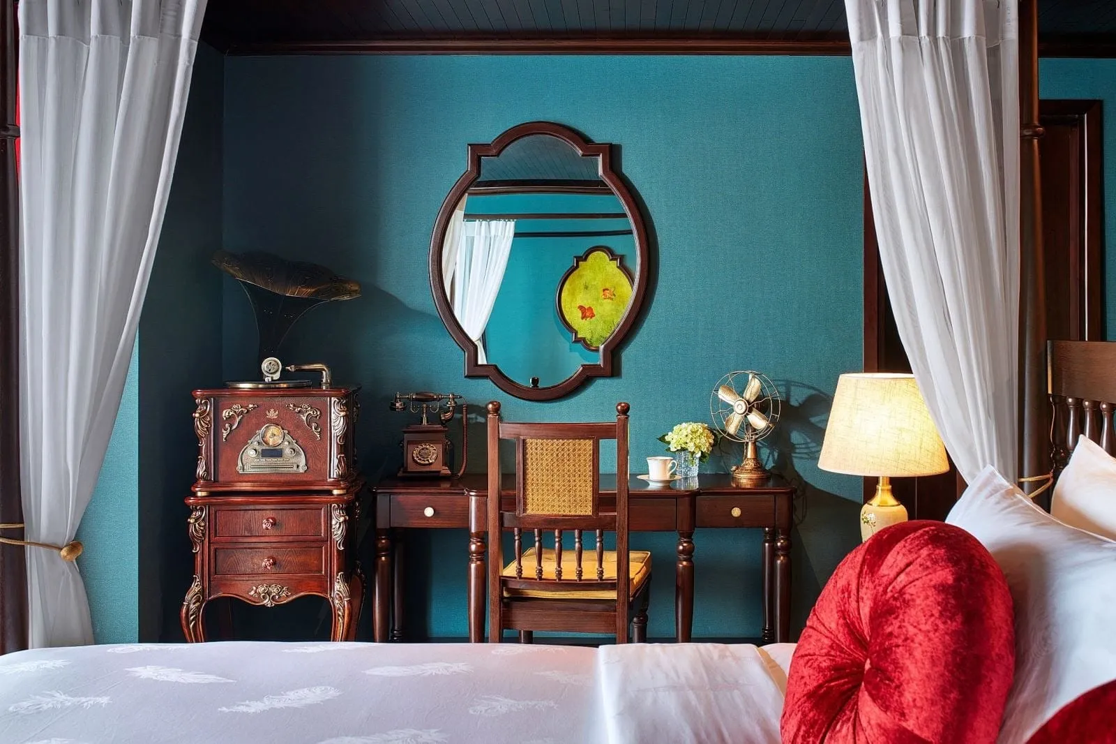 A vintage-style cabin with teal walls, a four-poster bed with white curtains, a dark wood desk with chair, gramophone cabinet, antique telephone, oval mirror, fan, lamp and a red velvet cushion.