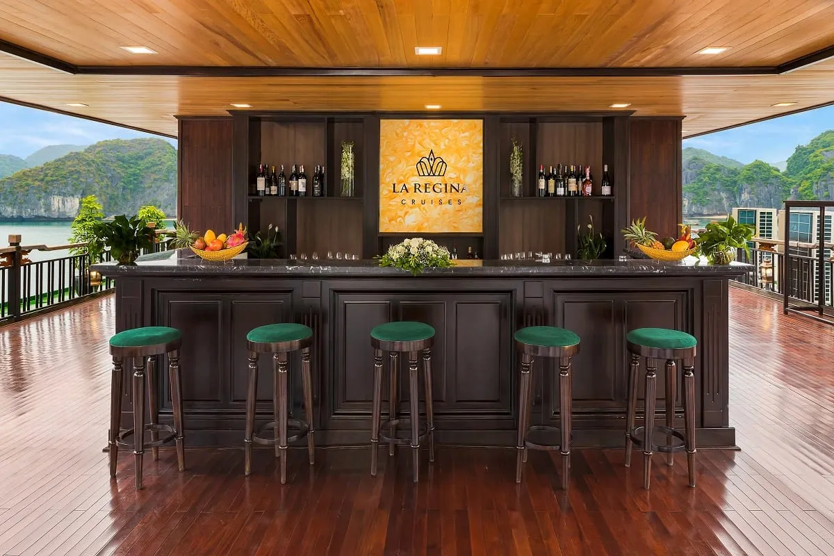 A wooden bar with green stools on a boat deck, with a backlit sign reading 'LA REGINA CRUISES' mounted on the wall, overlooking karst limestone mountains and calm water.
