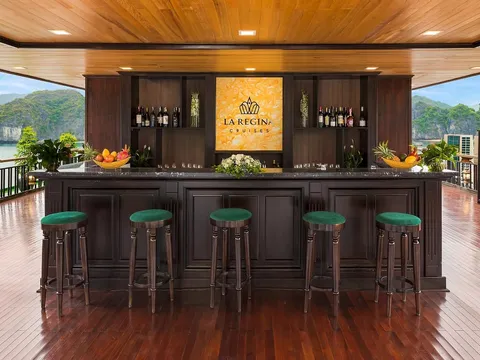 A wooden bar with green stools on a boat deck, with a backlit sign reading 'LA REGINA CRUISES' mounted on the wall, overlooking karst limestone mountains and calm water.