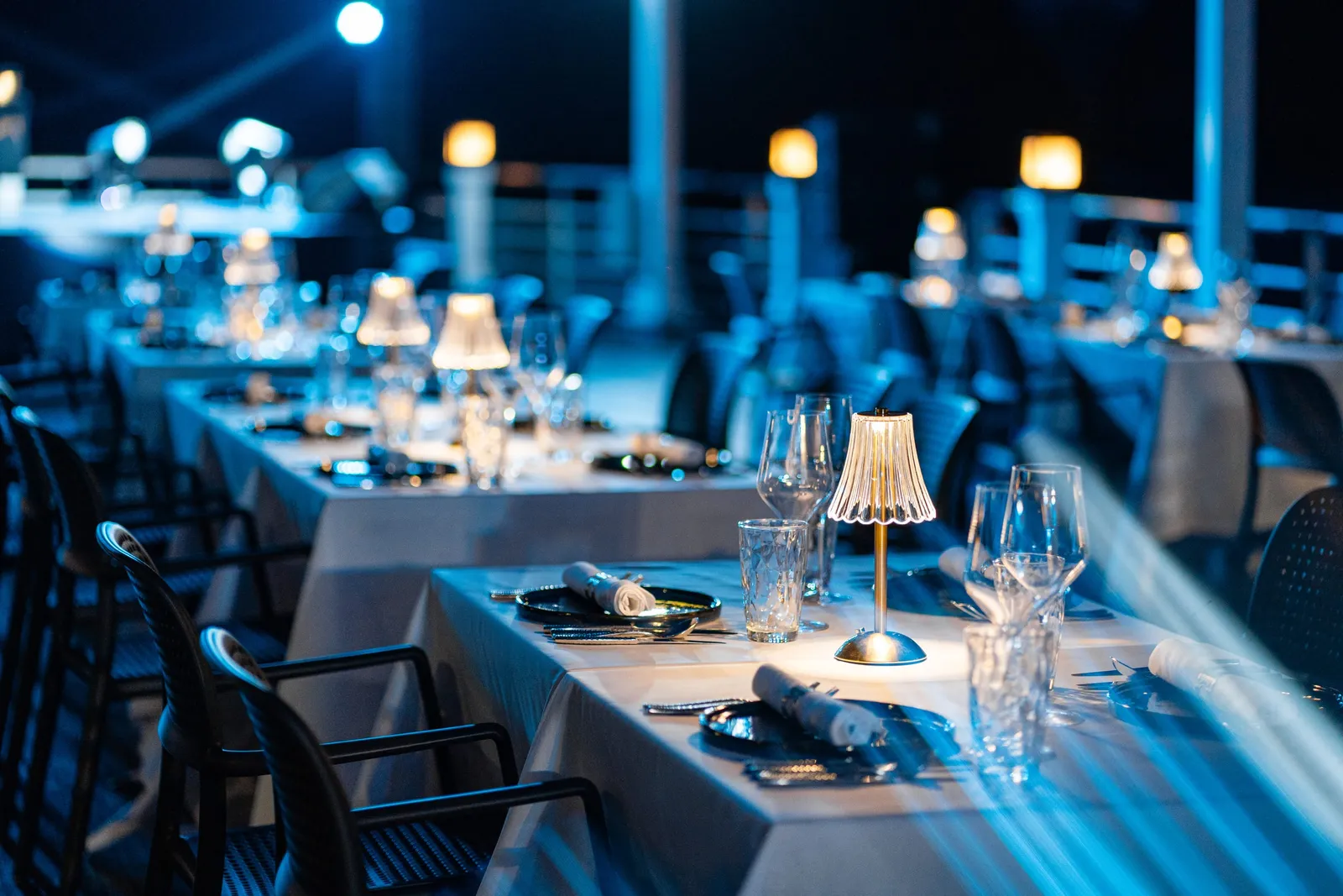 An evening dining area on a boat deck with set tables, small lamps, wine glasses, and chairs arranged in rows facing dark water.