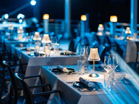 An evening dining area on a boat deck with set tables, small lamps, wine glasses, and chairs arranged in rows facing dark water.