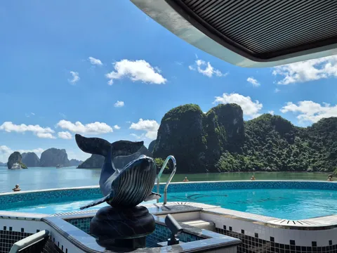 A tiled infinity pool with a whale sculpture on a boat's upper deck, overlooking calm water and forested limestone karsts under a blue sky, with a distant white cruise boat.