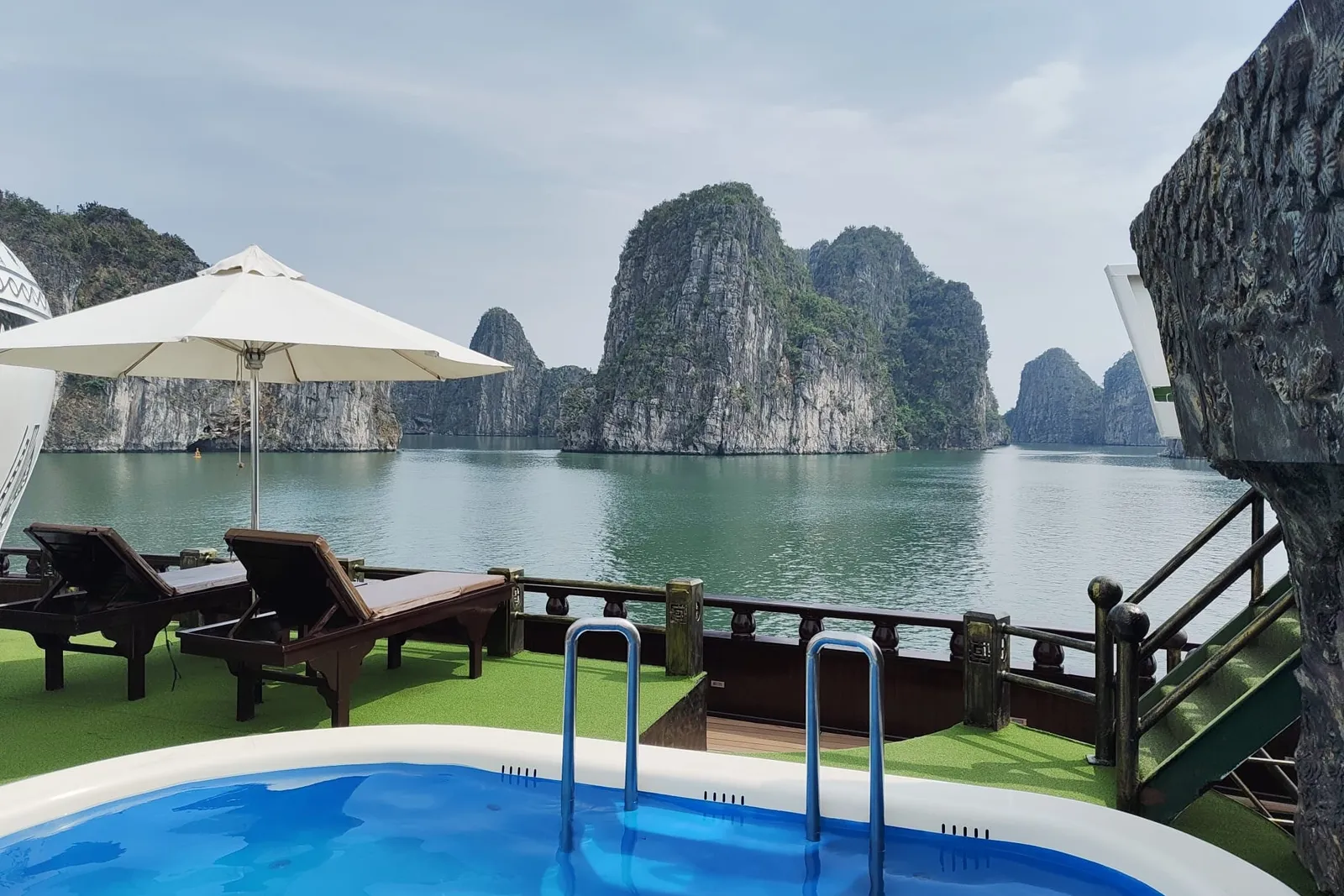 Deck pool with sun loungers under a white umbrella, the rail and open bay just beyond