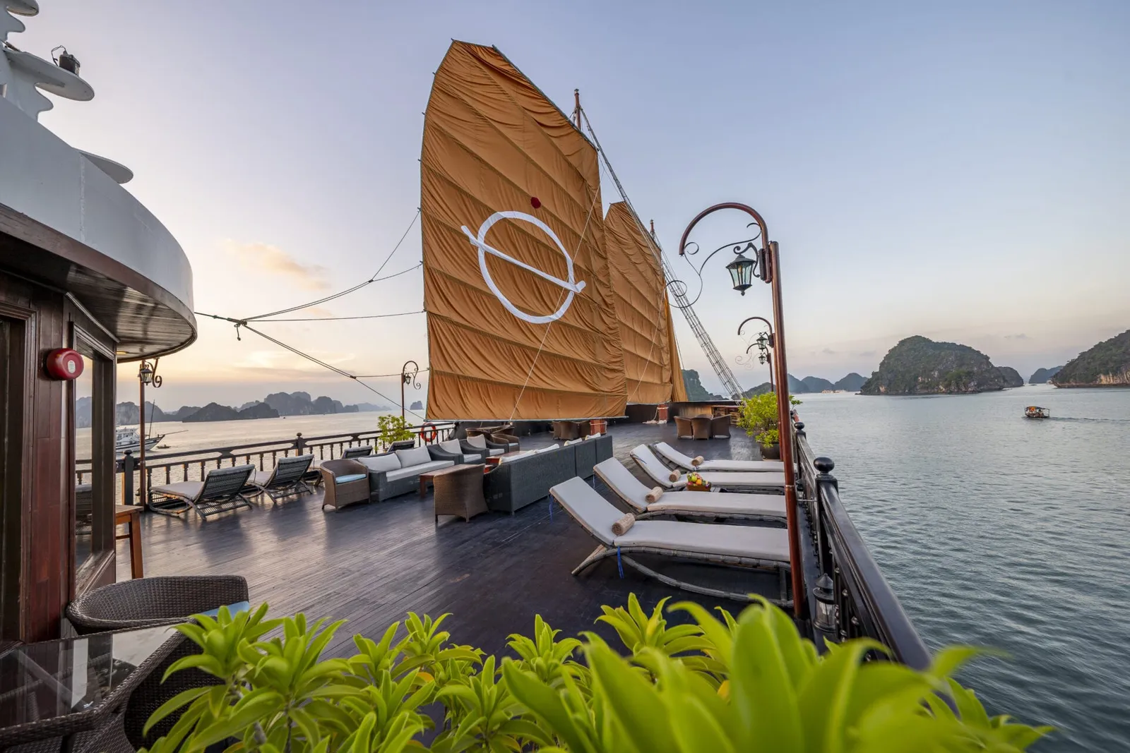 A wooden deck with lounge chairs and traditional orange sails featuring a white logo, overlooking limestone karst islands at sunset.