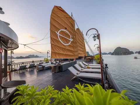 A wooden deck with lounge chairs and traditional orange sails featuring a white logo, overlooking limestone karst islands at sunset.
