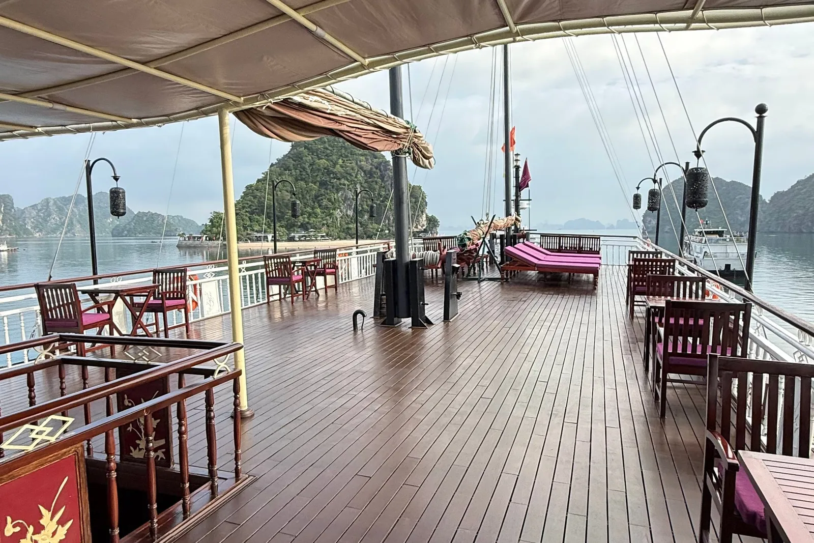 A wet wooden upper deck under a tan canopy with dark wood chairs, magenta cushions and loungers, lanterns on posts, a furled sail and rigging, and limestone karst islands in the bay beyond.