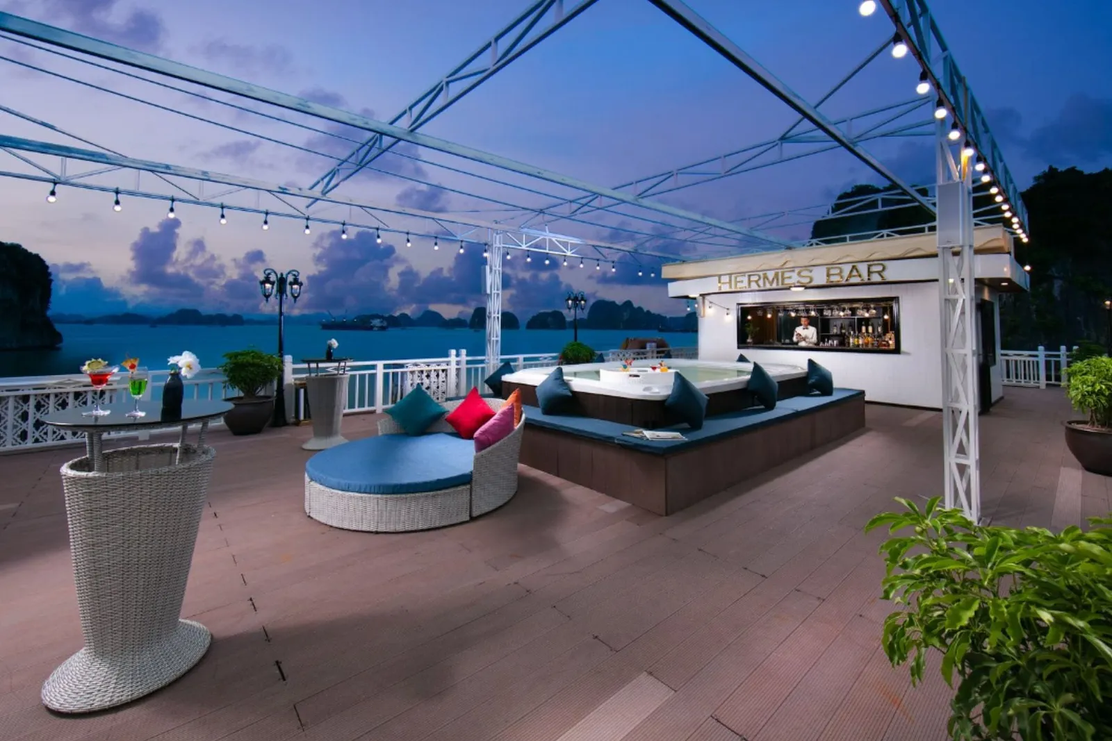 A cruise ship's outdoor deck at dusk with lounge seating, string lights, a bar labeled 'HERMES BAR', and limestone karsts visible across the water.