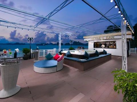 A cruise ship's outdoor deck at dusk with lounge seating, string lights, a bar labeled 'HERMES BAR', and limestone karsts visible across the water.