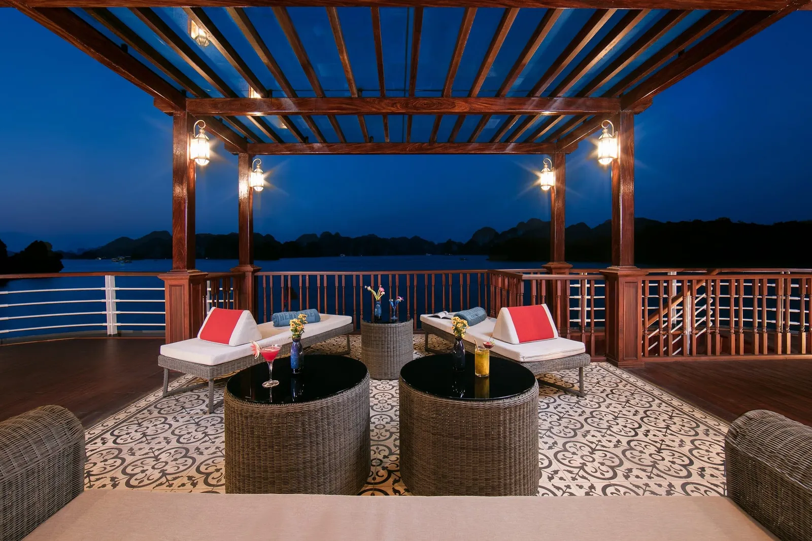 A wooden deck with lounge chairs, wicker tables holding champagne and flowers, under a pergola at dusk with limestone karsts visible across the water.