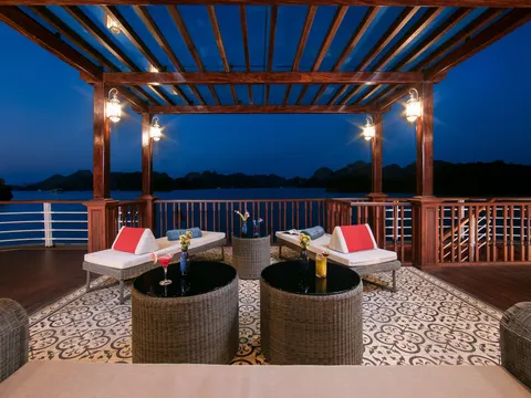 A wooden deck with lounge chairs, wicker tables holding champagne and flowers, under a pergola at dusk with limestone karsts visible across the water.