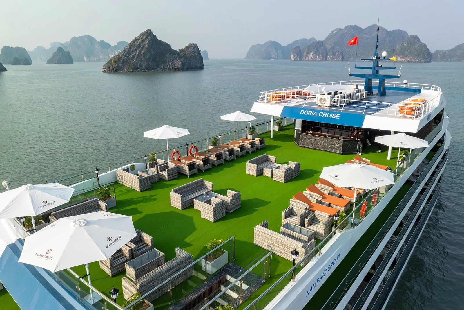 Aerial view of a cruise ship's top sun deck with green artificial turf, wicker lounge furniture, white Doria Cruise umbrellas and a bar, with limestone karsts rising from the bay behind.