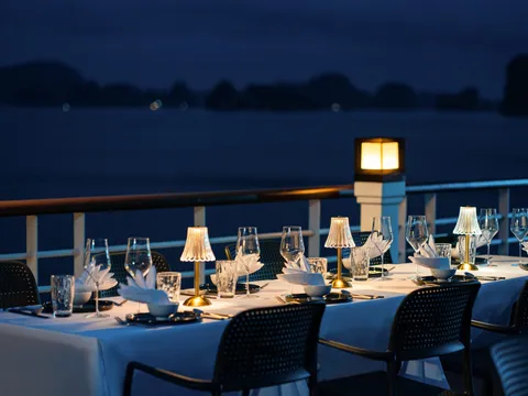 A formal dining table set with white linens, wine glasses, and small lamps on an outdoor deck at dusk with limestone karst formations visible across dark water in the background.