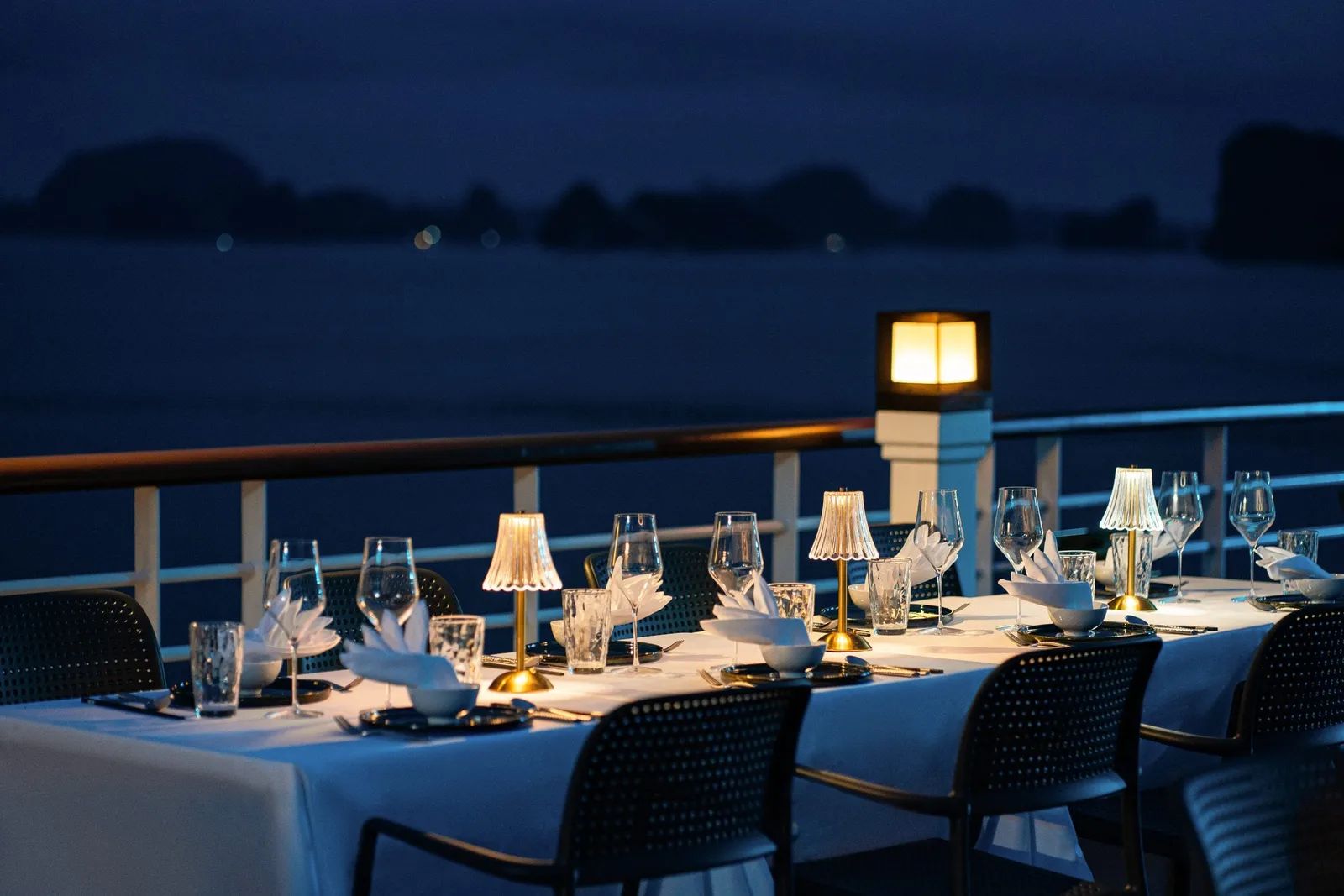 A formal dining table set with white linens, wine glasses, and small lamps on an outdoor deck at dusk with limestone karst formations visible across dark water in the background.