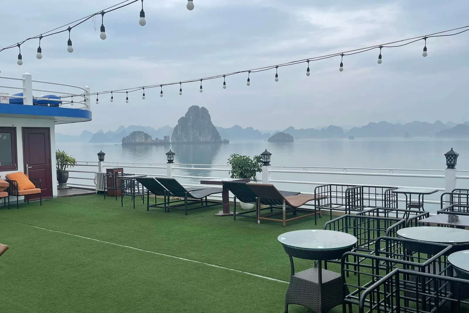 Sundeck laid with green matting, loungers and cafe tables under a run of string lights, karst beyond the rail