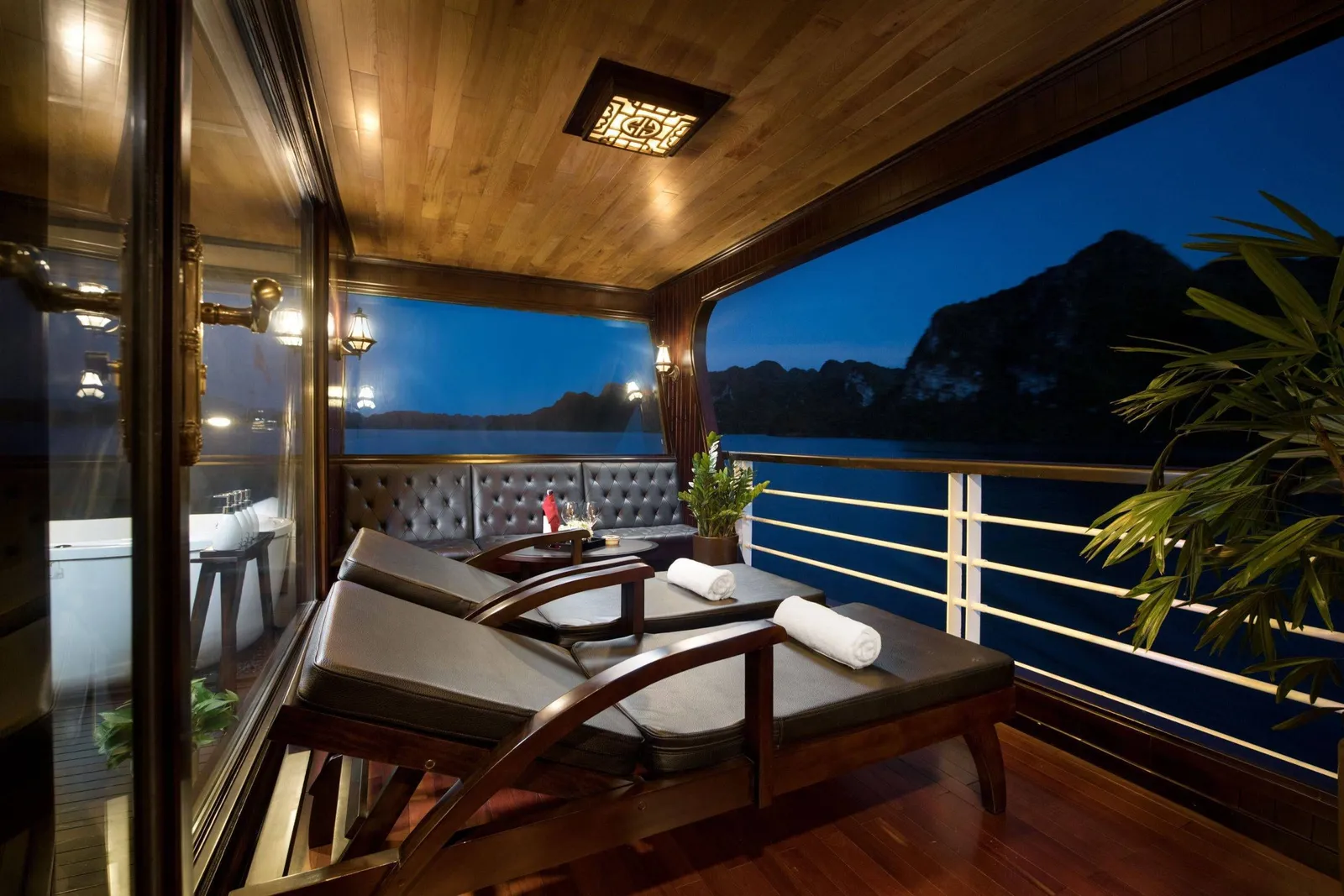 A covered private balcony on a boat at dusk with wooden loungers, rolled white towels, a tufted bench, a bathtub behind glass, and limestone karst silhouettes across the water.