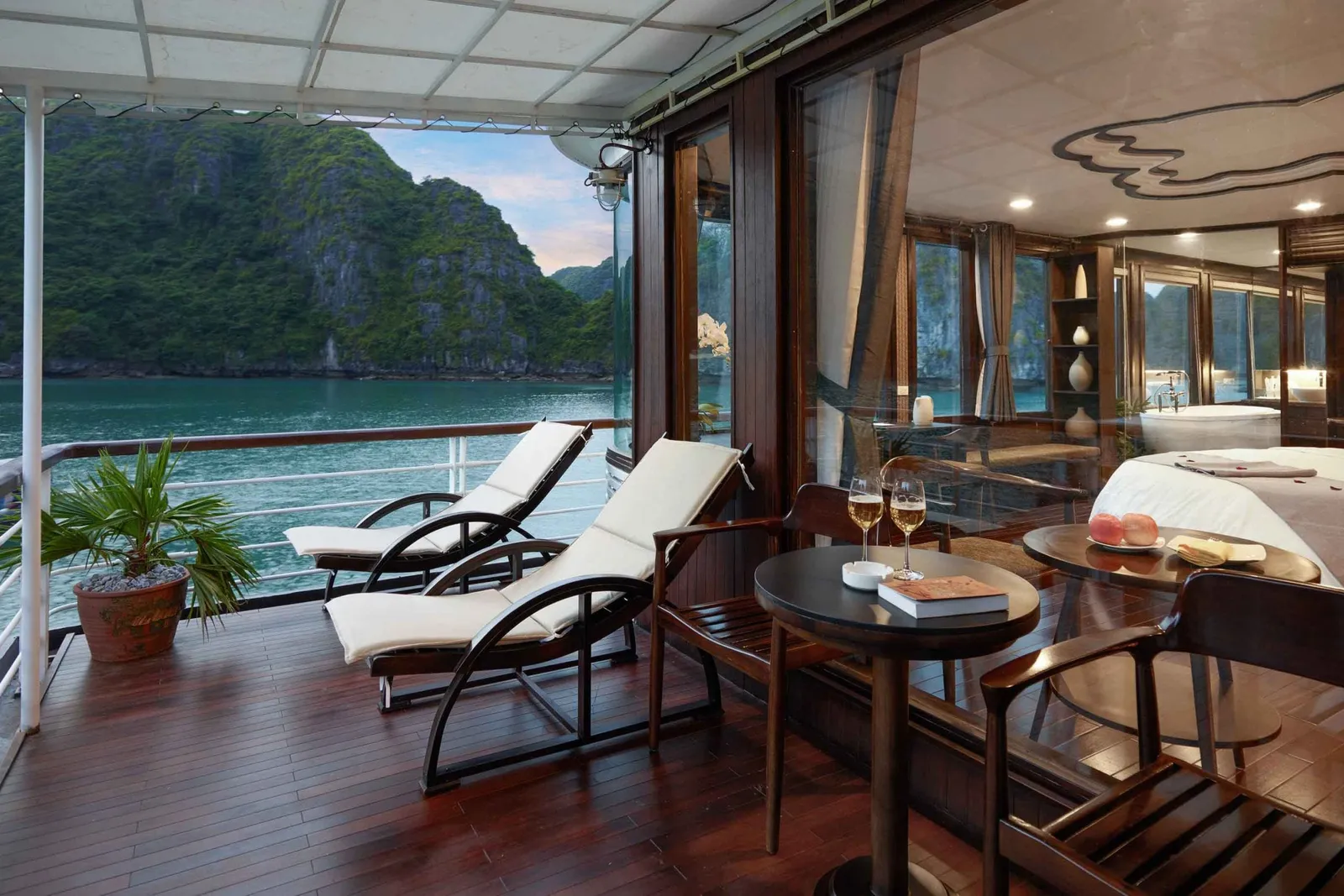 A wooden deck terrace with lounge chairs and a small table opens to water views of limestone karst islands through large glass doors leading to an interior cabin space.