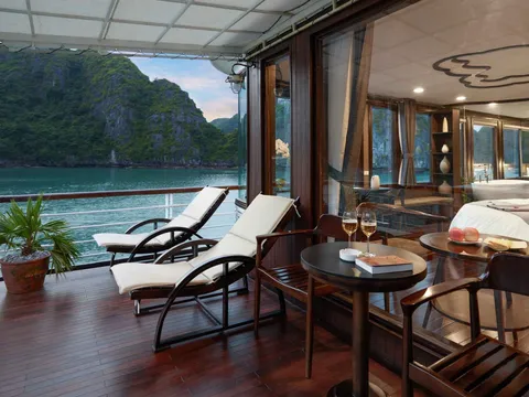 A wooden deck terrace with lounge chairs and a small table opens to water views of limestone karst islands through large glass doors leading to an interior cabin space.