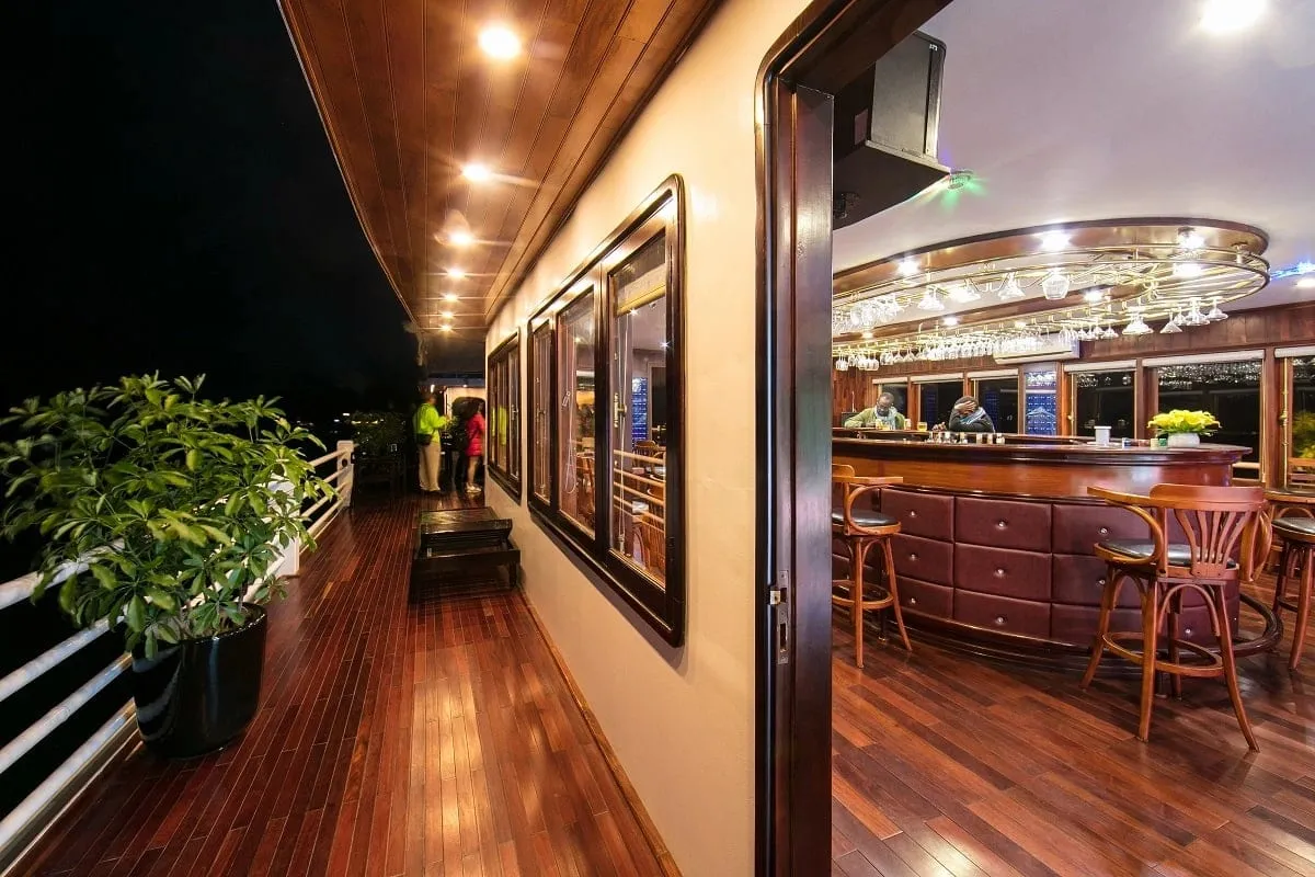 A night view of a polished wooden side deck with a white railing and a potted plant, next to an open door showing a curved bar with stools and hanging glasses, with a few people standing further along the deck.