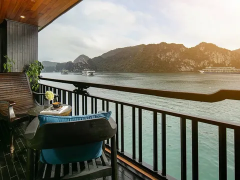 A wooden deck balcony with chairs and table overlooking calm water with limestone karst islands and cruise boats in the distance.