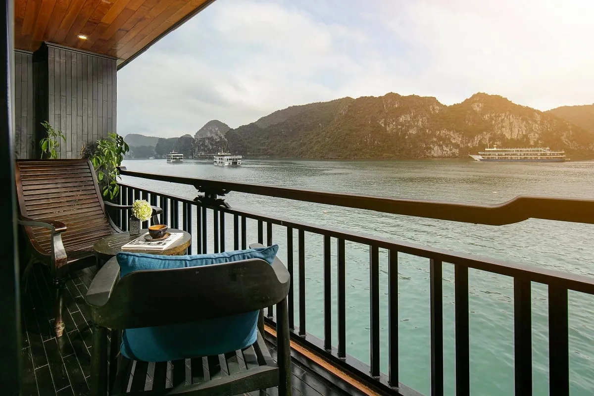 A wooden deck balcony with chairs and table overlooking calm water with limestone karst islands and cruise boats in the distance.