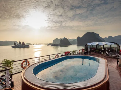 A cruise boat deck with a swimming pool and covered dining area during sunrise or sunset, with limestone karst islands and other cruise boats visible in the water.
