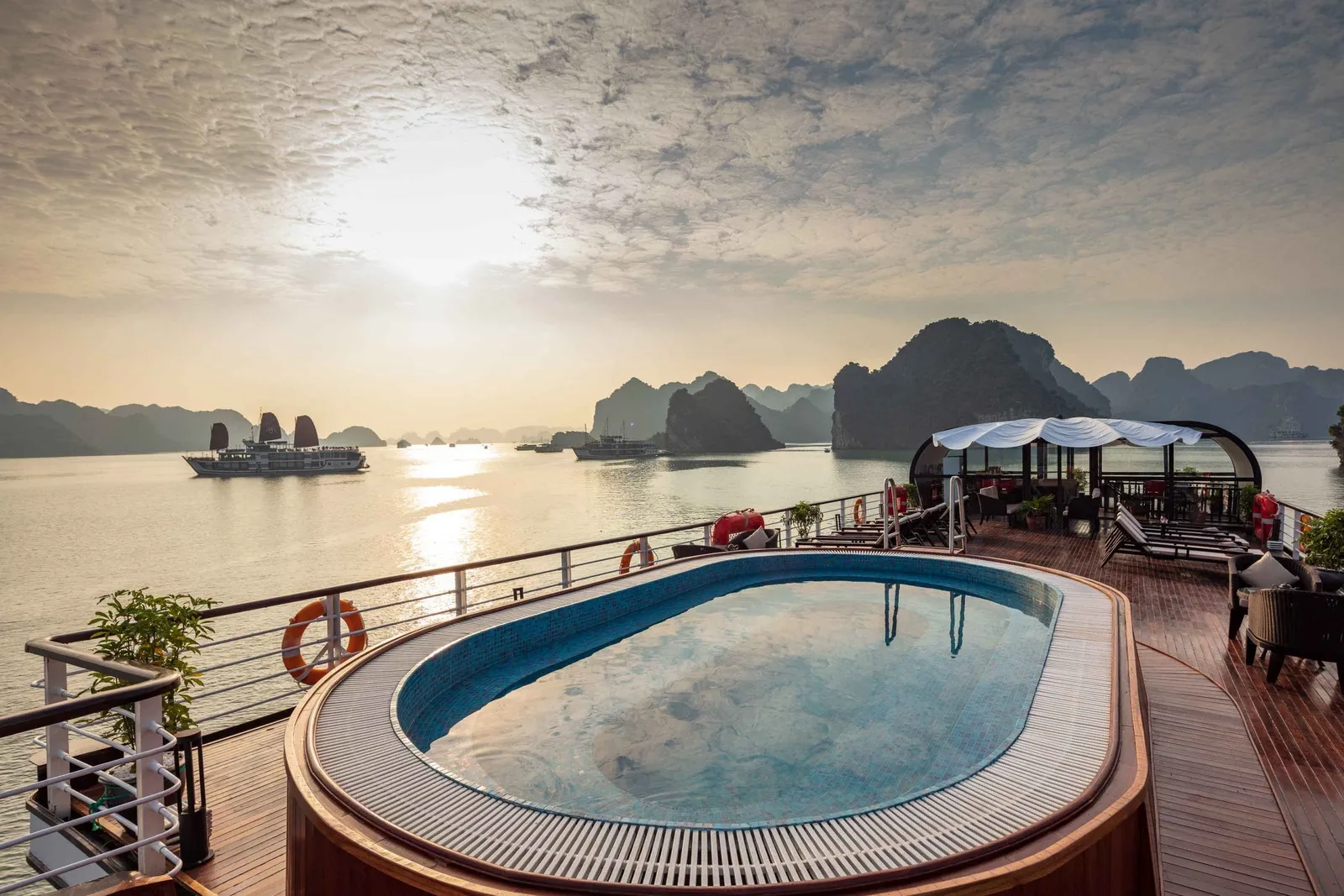 A cruise boat deck with a swimming pool and covered dining area during sunrise or sunset, with limestone karst islands and other cruise boats visible in the water.