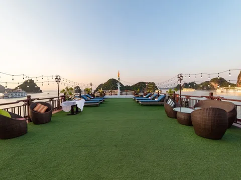 A sun deck covered in artificial grass with wicker chairs, blue loungers, a set table and string lights, overlooking calm water, limestone karst islands and other boats at dusk.