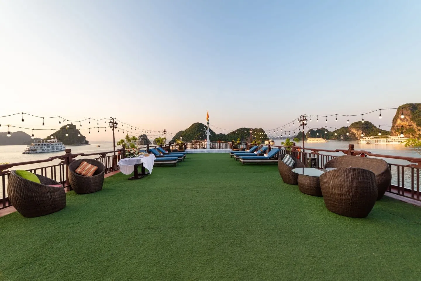 A sun deck covered in artificial grass with wicker chairs, blue loungers, a set table and string lights, overlooking calm water, limestone karst islands and other boats at dusk.