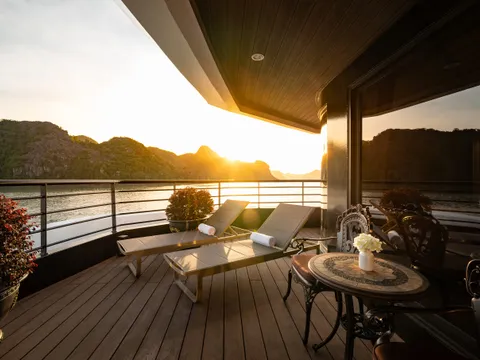 A wooden deck with sun loungers and furniture overlooking calm water with limestone karst formations at sunset.