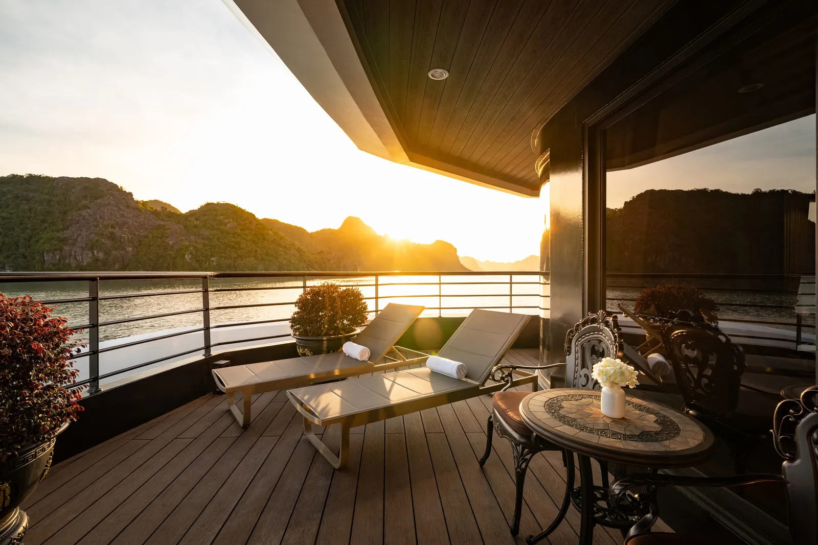 A wooden deck with sun loungers and furniture overlooking calm water with limestone karst formations at sunset.