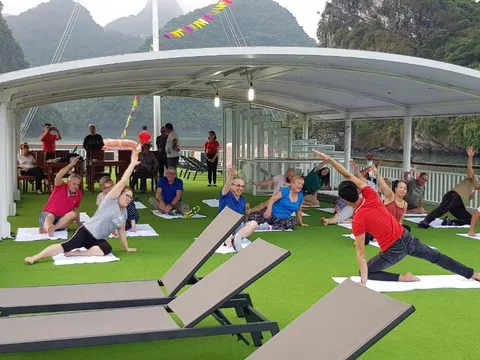 People doing yoga on white mats on a green artificial turf deck under a white canopy with dramatic karst limestone mountains and calm water in the background.
