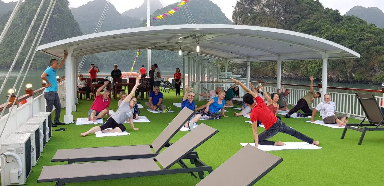 People doing yoga on white mats on a green artificial turf deck under a white canopy with dramatic karst limestone mountains and calm water in the background.