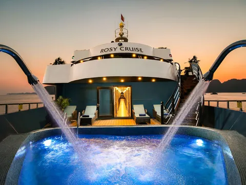 Stern jacuzzi lit blue after dark, four arcs of water crossing above it, the words Rosy Cruise on the deck behind