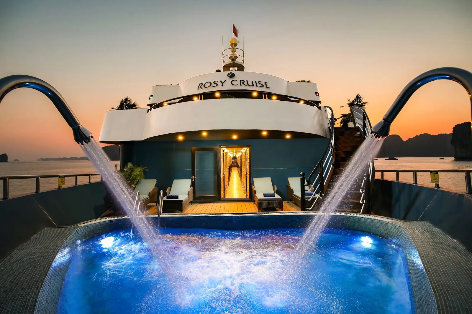 Stern jacuzzi lit blue after dark, four arcs of water crossing above it, the words Rosy Cruise on the deck behind