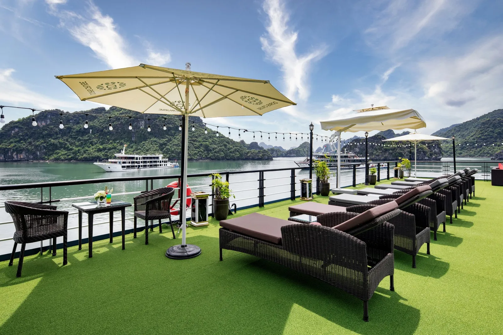 A sundeck with green artificial turf, dark wicker furniture, yellow umbrellas with printed text, and limestone karsts visible across the water with another cruise boat in the distance.