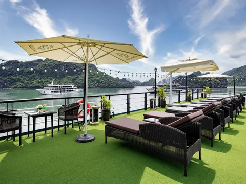 A sundeck with green artificial turf, dark wicker furniture, yellow umbrellas with printed text, and limestone karsts visible across the water with another cruise boat in the distance.