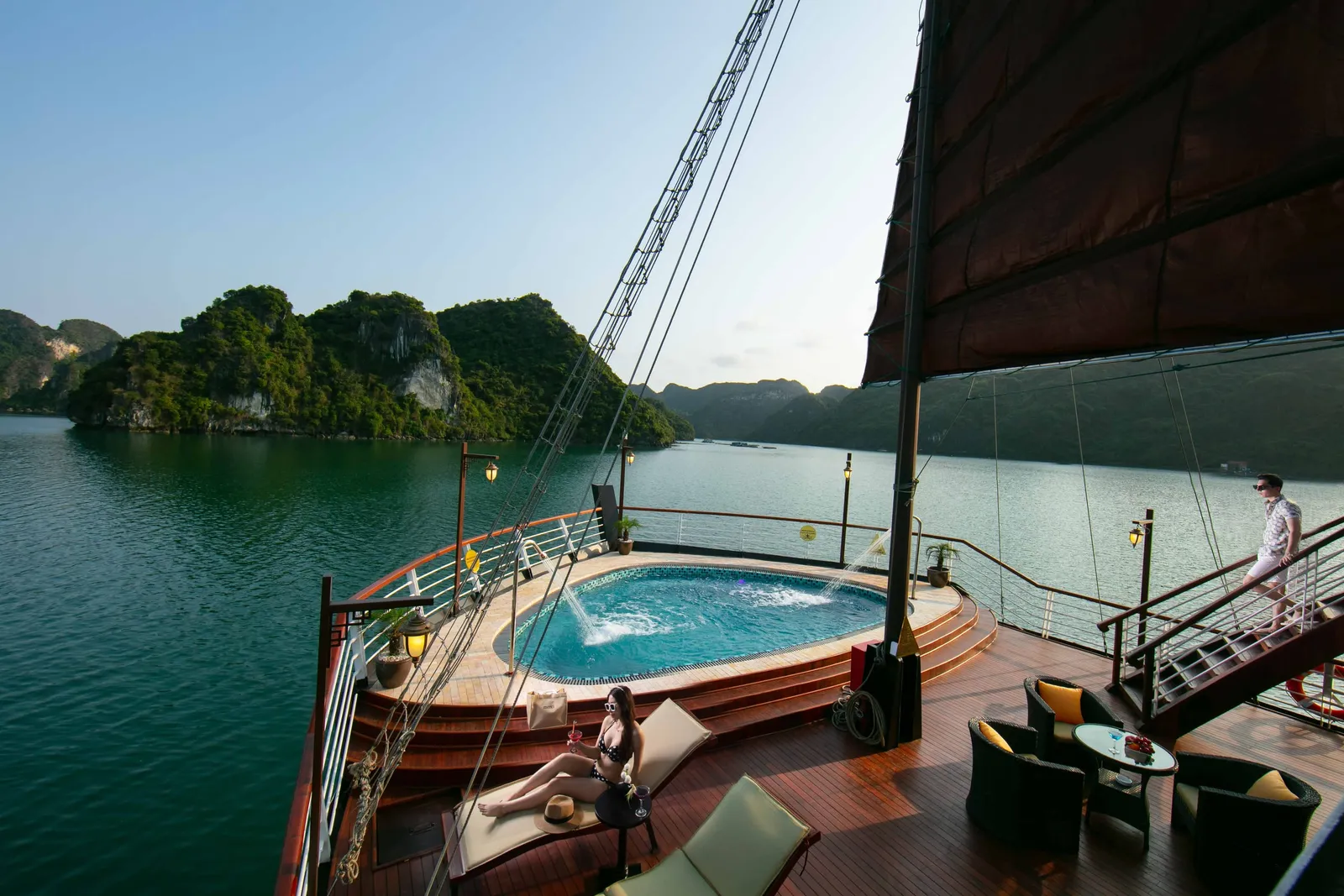 A wooden deck with a circular jacuzzi, lounge furniture, and staff member, with limestone karst islands in calm water behind.