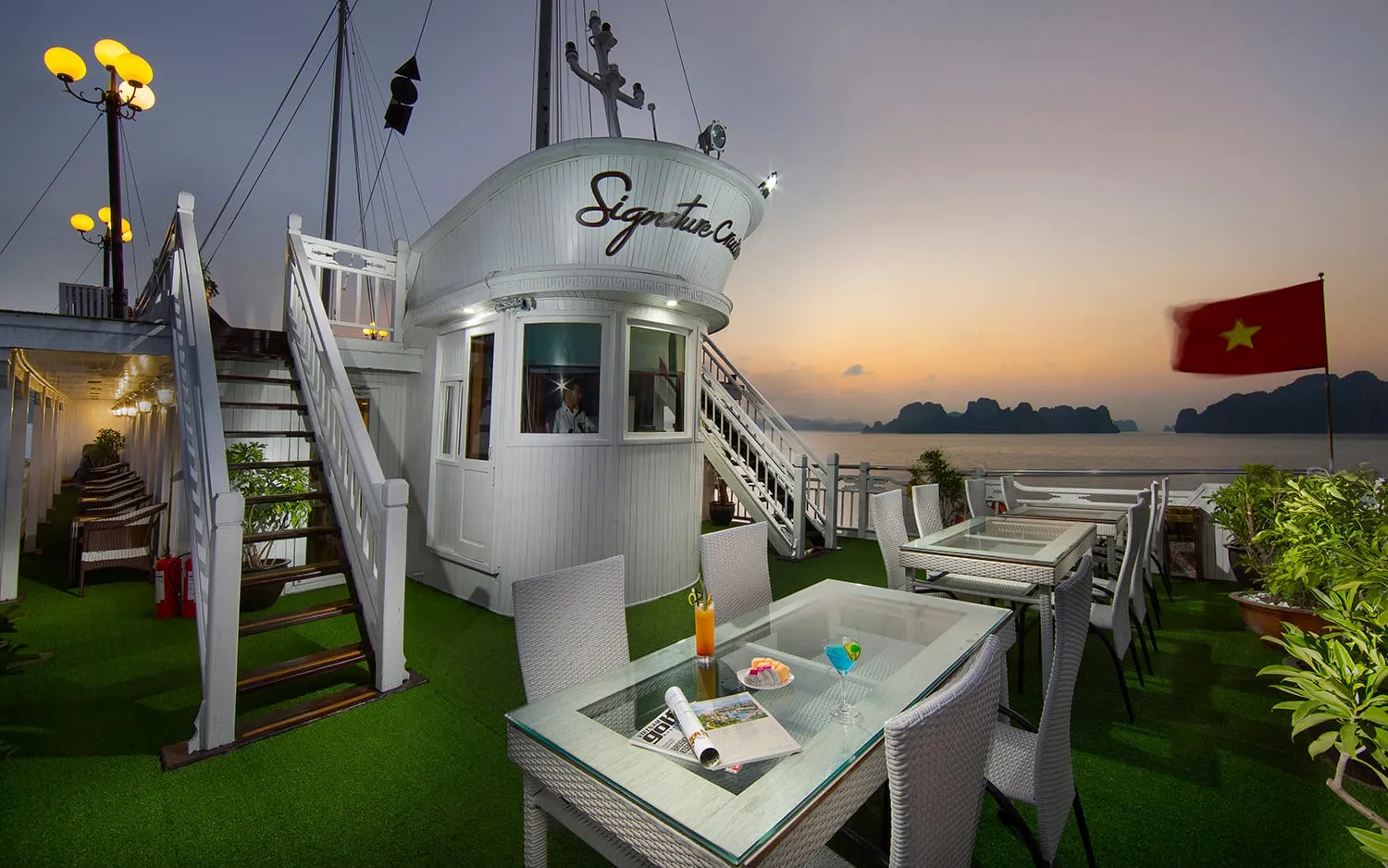 An outdoor deck on a cruise boat with wicker dining furniture on artificial grass, a white wheelhouse bearing the name 'Signature Cruise', a Vietnamese flag, and karst islands visible across the water at sunset.