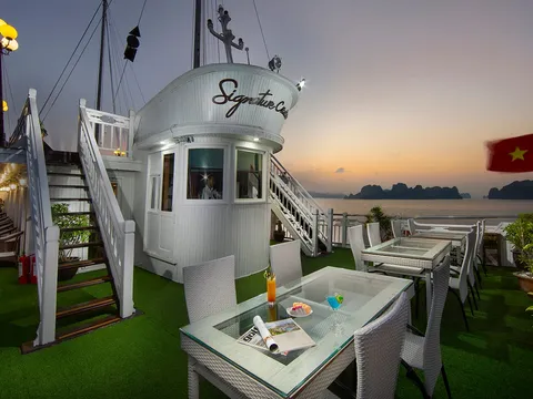 An outdoor deck on a cruise boat with wicker dining furniture on artificial grass, a white wheelhouse bearing the name 'Signature Cruise', a Vietnamese flag, and karst islands visible across the water at sunset.