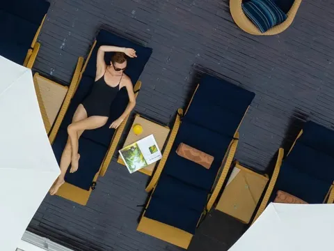 Overhead view of a woman in black swimwear relaxing on a blue deck lounger with wooden armrests, surrounded by other empty loungers on a dark wooden deck.