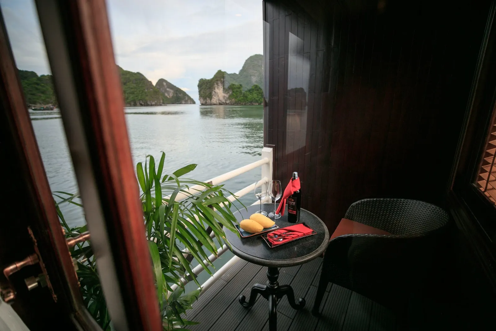 A private balcony on a wooden boat with a small round table holding wine, glasses and sliced mango, a wicker chair and a potted palm, overlooking calm water and limestone karst islands.