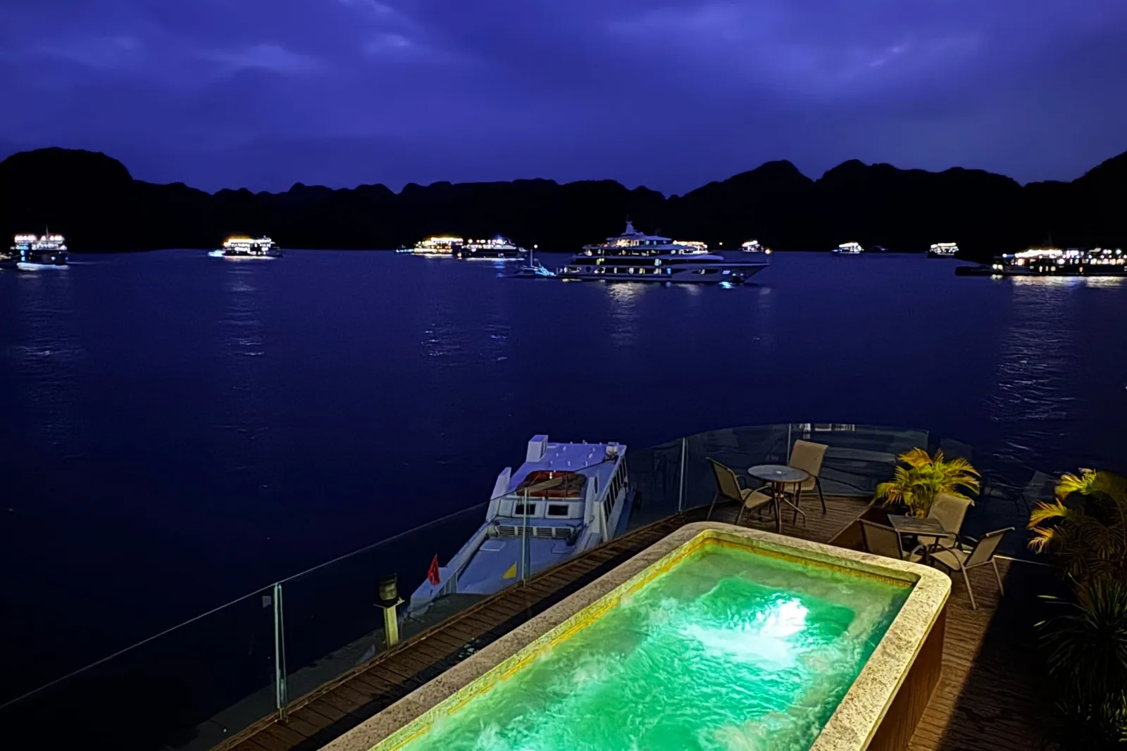 Deck pool lit green after dark, chairs set around it and the lights of other boats out on the water